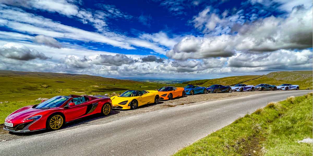 Eight McLaren Supercars parked behind each other along a countryside road underneath a blue and cloudy sky