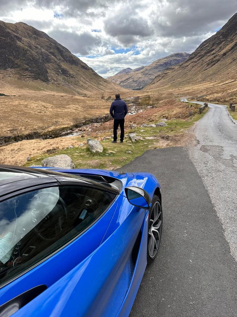 Wayne from Waynes World Of Wheels Supercar Club standing in front of a Mclaren supercar looking down Glencoe valley in Scotland where Skyfall James Bond was filmed