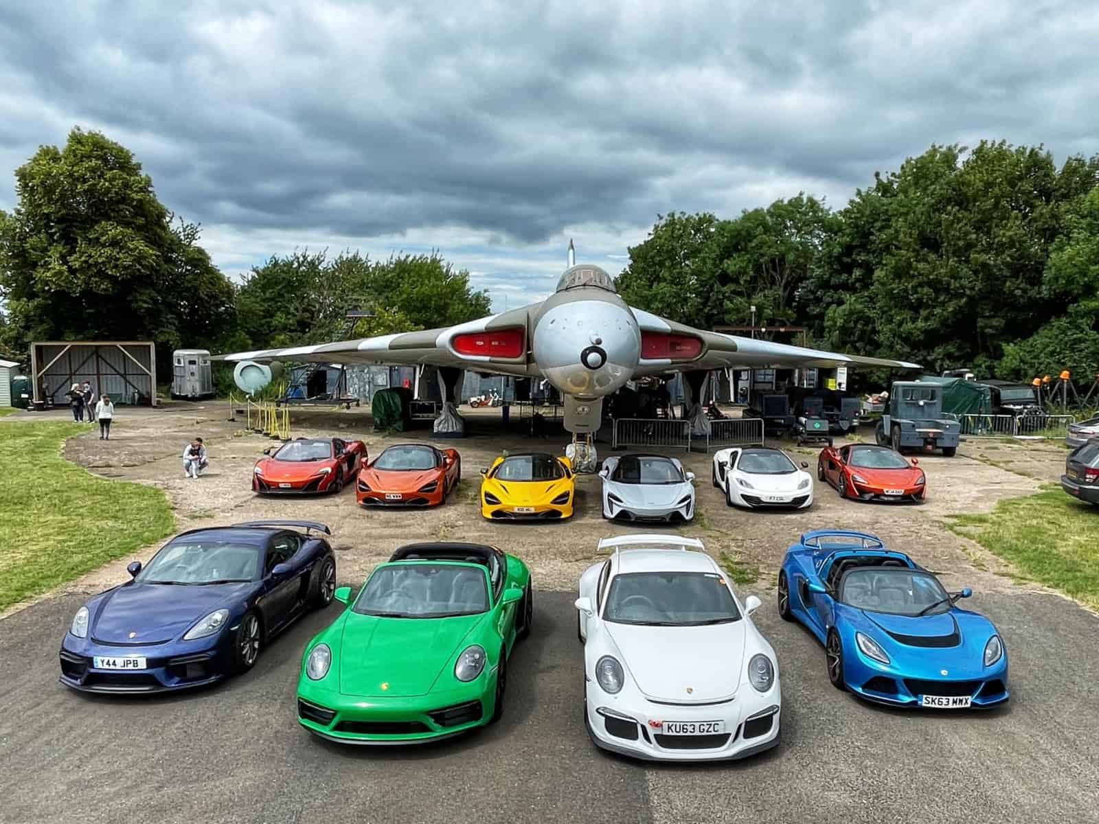 A mixture of Supercars and McLaren Supercars parked on the runway underneath a Vulcan Bomber Airplane as part of the WWOW Vulcan Bomber Day drive