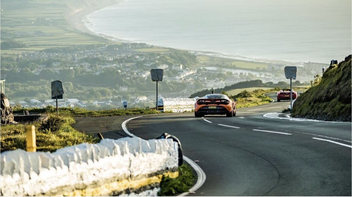 image of an Orange Mclaren Supercar driving on a sunny morning on the open roads of the Isle Of Man while on a Waynes World Of Wheels Tour